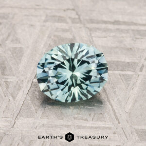 A 0.97-carat Montana Sapphire, faceted in a light blue oval, rests on a textured gray surface above the "EARTH'S TREASURY" name and hexagonal logo.