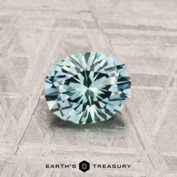 0.97-Carat Montana Sapphire A 0.97-carat Montana Sapphire, faceted in a light blue oval, rests on a textured gray surface above the "EARTH'S TREASURY" name and hexagonal logo.