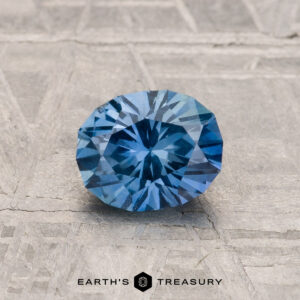 The 1.02-Carat Montana Sapphire, round and faceted, is shown on a textured gray surface with "EARTH'S TREASURY" printed below.