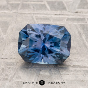 A 2.33-Carat Montana Sapphire, rectangular and faceted in vibrant violet-blue, is displayed on a textured surface with "EARTH'S TREASURY" shown at the bottom.