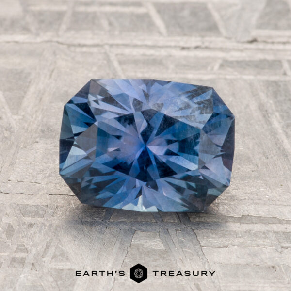 2.33-Carat Montana Sapphire A 2.33-Carat Montana Sapphire, rectangular and faceted in vibrant violet-blue, is displayed on a textured surface with "EARTH'S TREASURY" shown at the bottom.