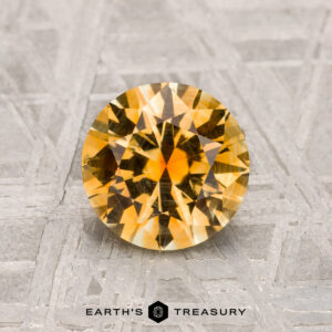A vibrant yellow, round faceted 1.52-carat Montana Sapphire is displayed on a textured gray surface with "EARTH'S TREASURY" at the bottom.