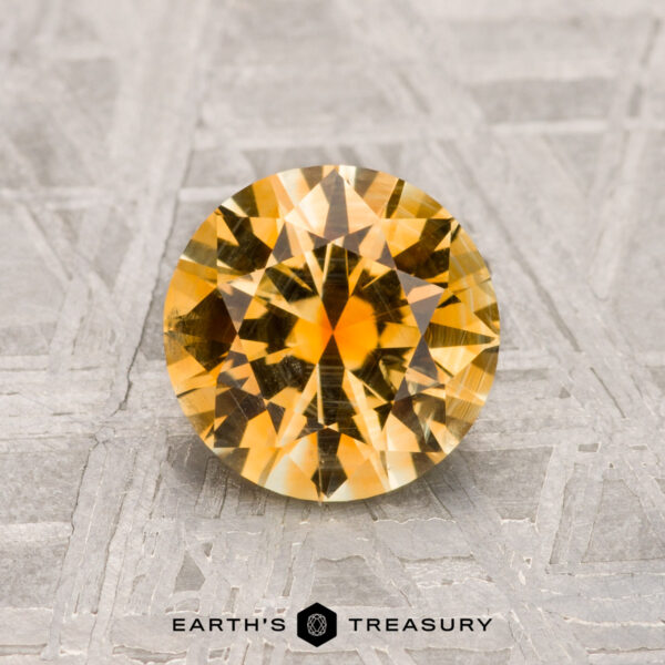 A vibrant yellow, round faceted 1.52-carat Montana Sapphire is displayed on a textured gray surface with "EARTH'S TREASURY" at the bottom.