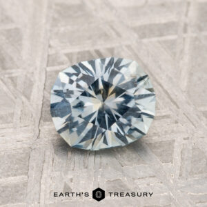 A 1.29-carat Montana Sapphire with a bluish-gray hue rests on a textured surface. The "EARTH'S TREASURY" logo and text are shown at the bottom.