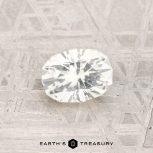 The 0.81-Carat Montana Sapphire, a faceted near colorless gemstone, is shown on a textured geometric surface with "EARTH'S TREASURY" displayed at the bottom.