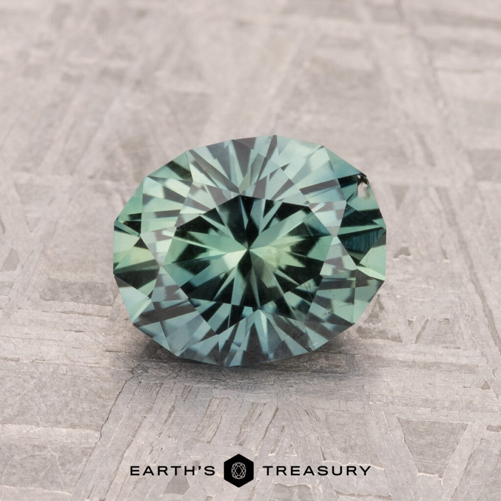 A 1.67-carat Montana sapphire, round and teal with faceted cuts, rests on a textured surface. Below it reads "Earth's Treasury.