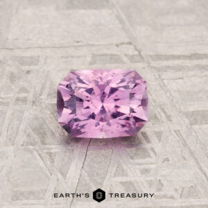 A 0.69-carat Montana Sapphire with a radiant cut is displayed on a textured gray surface, featuring the "Earth's Treasury" text at the bottom.