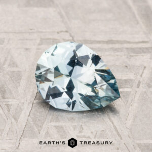 The 1.24-Carat Montana Sapphire, showcasing a light aqua blue hue, is positioned on a textured gray surface with the "Earth's Treasury" logo displayed below.