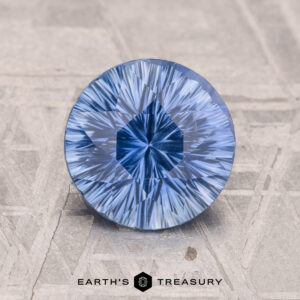 A round blue 2.10-carat Ceylon sapphire with a starburst pattern sits on a textured gray surface. "Earth's Treasury" logo at the bottom.