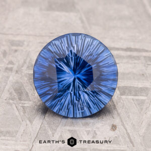 A 1.84-carat, brilliantly cut round Ceylon sapphire with intricate patterns is placed on a textured gray surface. "Earth's Treasury" is visible at the bottom.