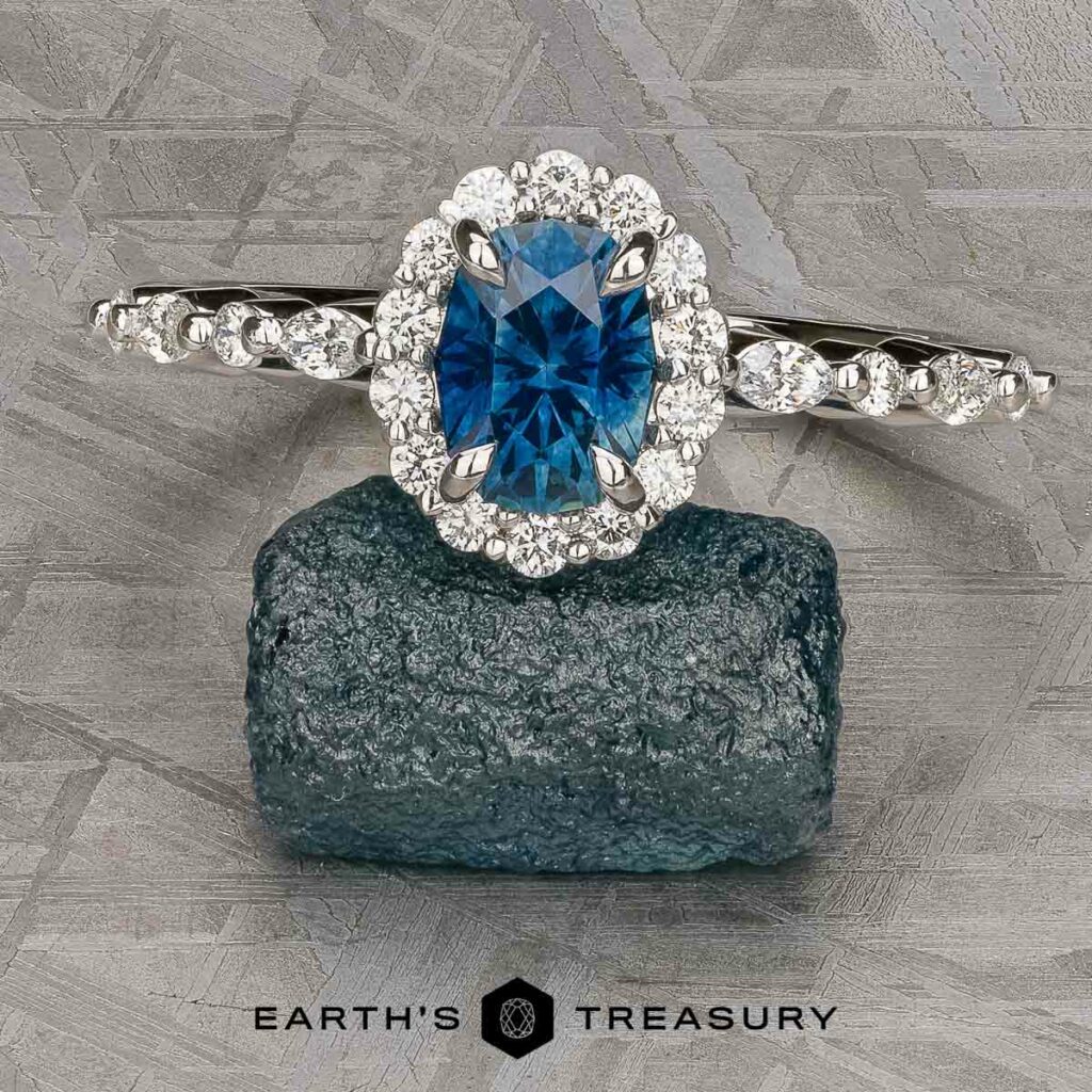 The Stacked from 56 images. Method=B (R=1,S=3) ring, featuring an oval blue gemstone with a halo of diamonds and a diamond-accented band, rests on a textured gray surface.