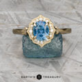 The "Ailette" ring in 14K yellow gold, featuring a 1.44-carat Montana sapphire surrounded by diamonds, displayed on blue crystal with the "Earth's Treasury" logo below.