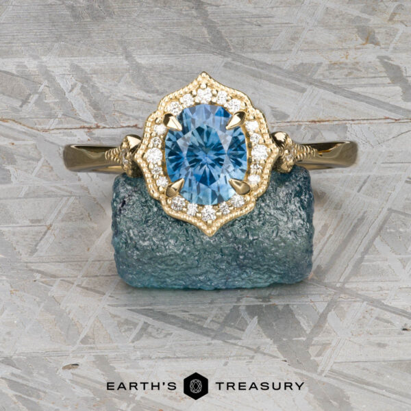 The "Ailette" ring in 14K yellow gold with 1.44-carat Montana sapphire The "Ailette" ring in 14K yellow gold, featuring a 1.44-carat Montana sapphire surrounded by diamonds, displayed on blue crystal with the "Earth's Treasury" logo below.
