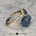 The "Ailette" ring in 14K yellow gold, featuring a 1.44-carat Montana sapphire and accent stones, rests on a rough blue mineral atop a textured surface. The text "Earth's Treasury" appears below.