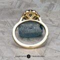 The "Ailette" ring in 14K yellow gold with a 1.44-carat Montana sapphire stands upright on a textured gray surface, its intricate design framing the rough blue stone above the Earth's Treasury logo.
