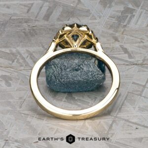 The "Ailette" ring in 14K yellow gold with a 1.44-carat Montana sapphire stands upright on a textured gray surface, its intricate design framing the rough blue stone above the Earth's Treasury logo.