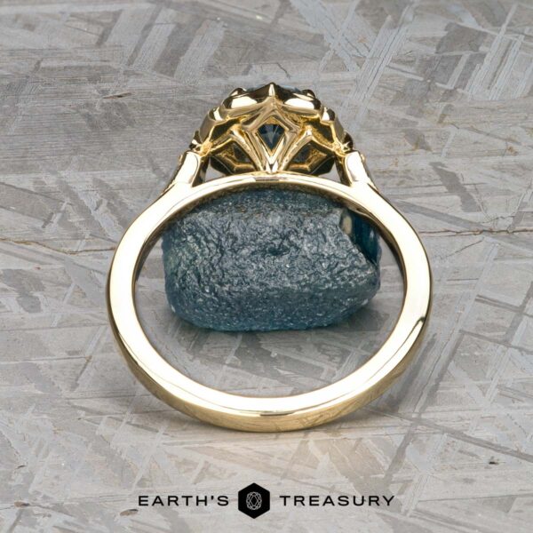 The "Ailette" ring in 14K yellow gold with 1.44-carat Montana sapphire The "Ailette" ring in 14K yellow gold with a 1.44-carat Montana sapphire stands upright on a textured gray surface, its intricate design framing the rough blue stone above the Earth's Treasury logo.