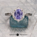 The "Ailette" ring in 18K white gold features a 1.57-carat oval Umba sapphire, surrounded by sparkling stones and showcased on a textured blue-green rock with the "Earth's Treasury" logo below.