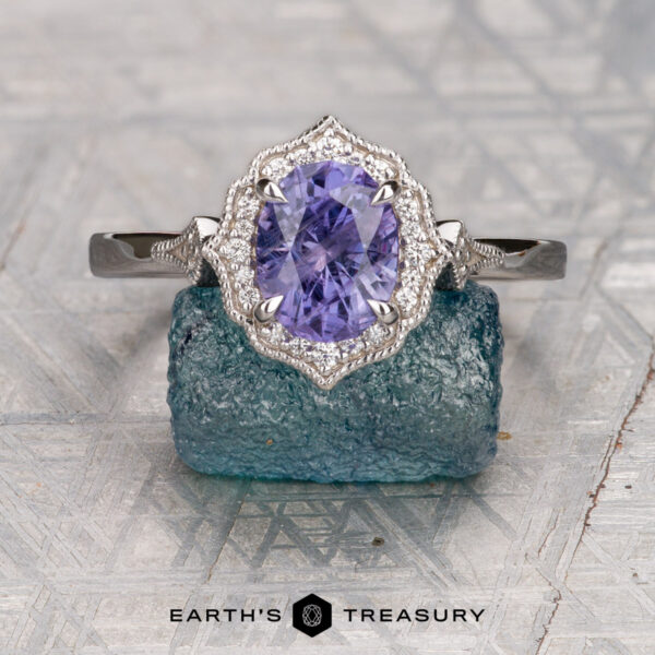 The "Ailette" ring in 18K white gold with 1.57-carat Umba sapphire The "Ailette" ring in 18K white gold features a 1.57-carat oval Umba sapphire, surrounded by sparkling stones and showcased on a textured blue-green rock with the "Earth's Treasury" logo below.