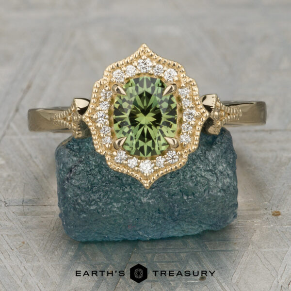 The "Ailette" ring in 14K yellow gold with 0.94-carat Australian sapphire The "Ailette" ring in 14K yellow gold features a 0.94-carat Australian sapphire center, accented by small diamonds and displayed on a rough blue stone surface. The text "EARTH'S TREASURY" appears at the bottom.