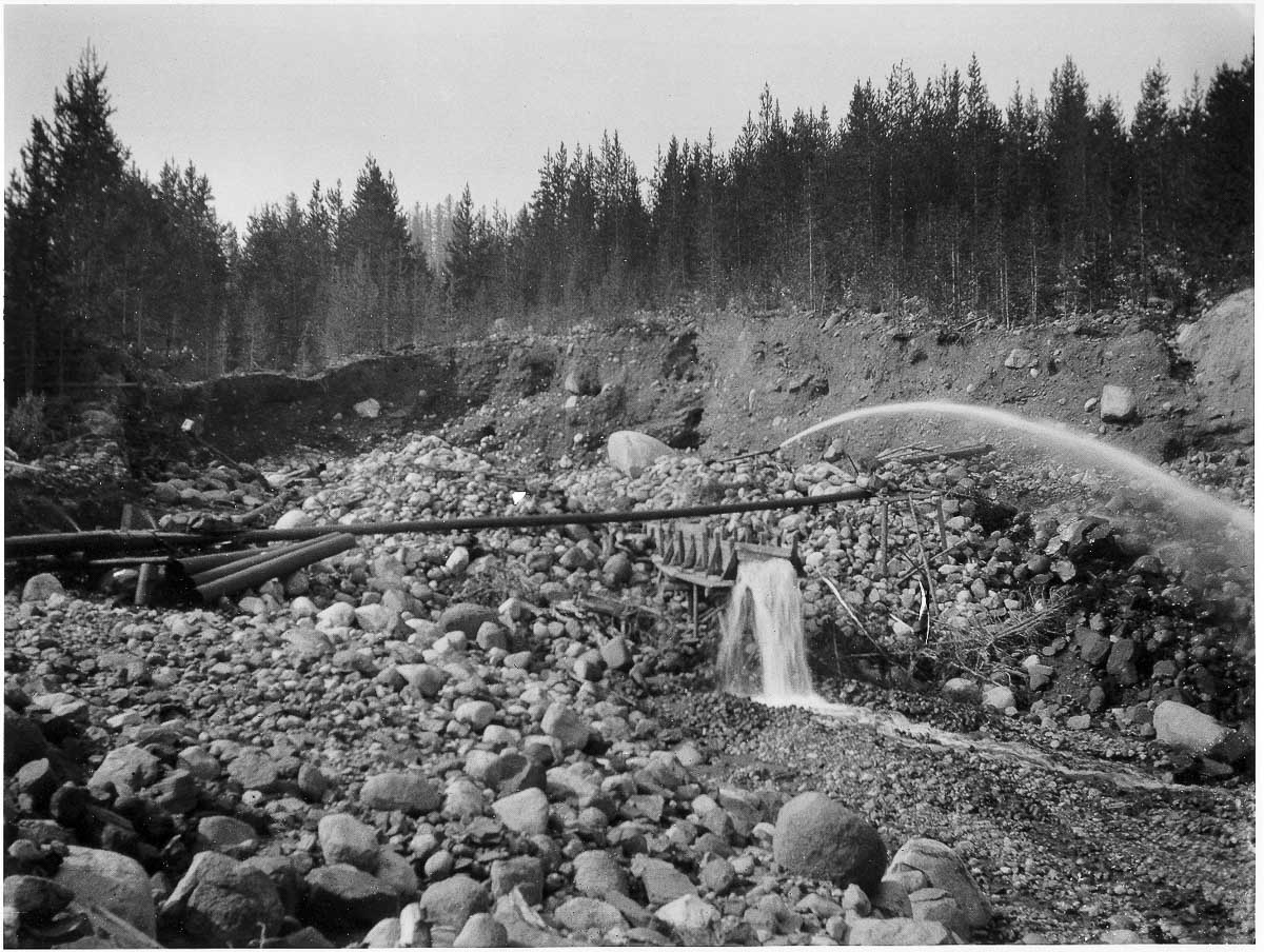 The Hydraulic mining-1200-BW channels water through pipes across a rocky landscape enveloped by forest.
