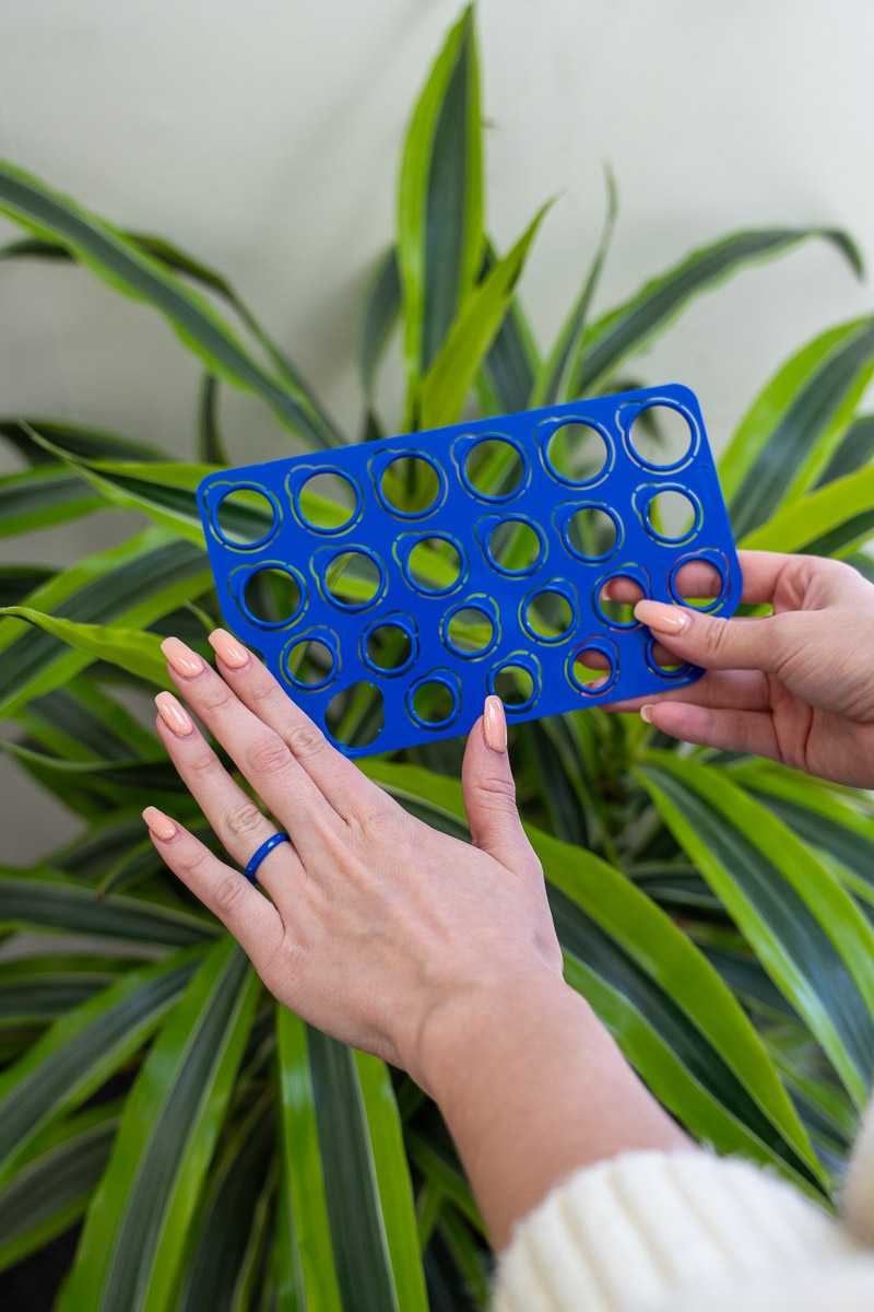 A person holds the Ring Sizer-1200-BW, a blue plastic stencil with circular cutouts, in front of green plant leaves.