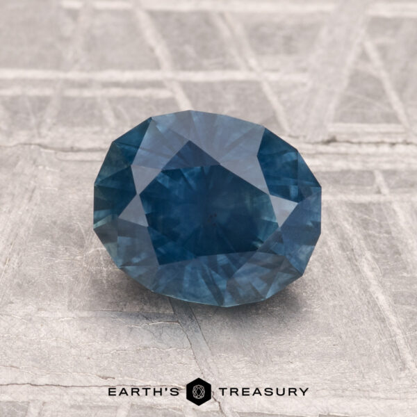 1.69-Carat Montana Sapphire (Heated) A 1.69-carat heated Montana sapphire with a blue hue is displayed on a textured light gray surface, featuring the "Earth's Treasury" logo at the bottom.