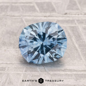 A round, faceted 1.93-carat heated Montana sapphire is showcased on a textured gray surface with "EARTH'S TREASURY" displayed at the bottom.