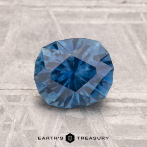 A 1.56-carat heated Montana sapphire with a blue faceted cut rests on a textured gray surface, "Earth's Treasury" written below.