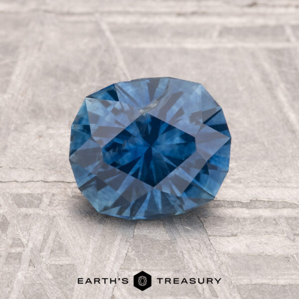 1.56-Carat Montana Sapphire (Heated) A 1.56-carat heated Montana sapphire with a blue faceted cut rests on a textured gray surface, "Earth's Treasury" written below.