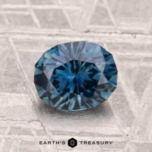 A round, faceted 1.58-carat Montana Sapphire (Heated) is displayed on a textured metallic surface with "EARTH'S TREASURY" at the bottom.