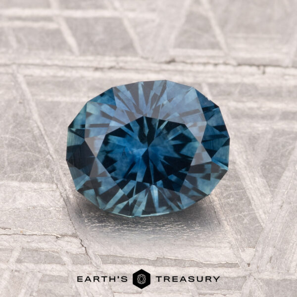 1.58-Carat Montana Sapphire (Heated) A round, faceted 1.58-carat Montana Sapphire (Heated) is displayed on a textured metallic surface with "EARTH'S TREASURY" at the bottom.