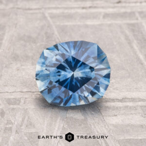 A 1.45-carat heated Montana sapphire with a brilliant cut is displayed on a textured gray surface, with "EARTH'S TREASURY" shown below.