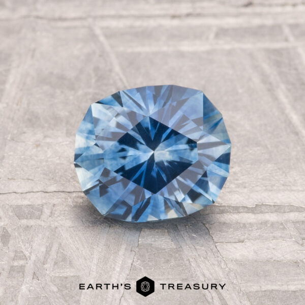 1.45-Carat Montana Sapphire (Heated) A 1.45-carat heated Montana sapphire with a brilliant cut is displayed on a textured gray surface, with "EARTH'S TREASURY" shown below.