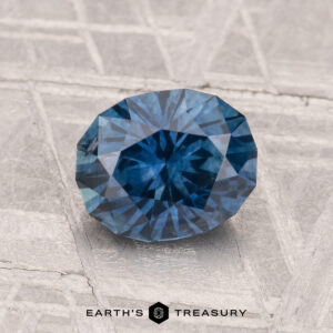 A faceted, oval-shaped 1.57-carat heated Montana sapphire shown on a textured gray surface, with the "Earth's Treasury" logo and text at the bottom.