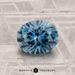 A 1.49-carat heated Montana sapphire, round and blue with faceted cuts, is shown on a textured gray surface with "Earth's Treasury" written below.