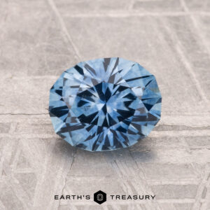 A 1.45-carat heated Montana sapphire with a round brilliant cut rests on a textured gray surface, featuring the text "Earth's Treasury" at the bottom.