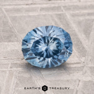 A 1.44-carat heated Montana Sapphire with a round brilliant cut is shown on a textured gray surface, with "EARTH'S TREASURY" written at the bottom.