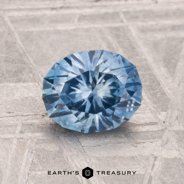 1.44-Carat Montana Sapphire (Heated) A 1.44-carat heated Montana Sapphire with a round brilliant cut is shown on a textured gray surface, with "EARTH'S TREASURY" written at the bottom.