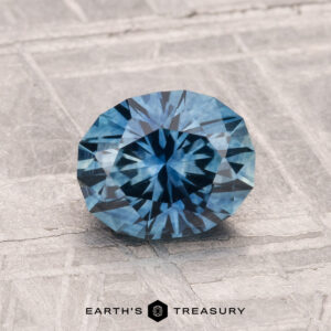 1.41-carat heated Montana sapphire, faceted oval and blue, displayed on a textured surface with the Earth’s Treasury logo at the bottom.