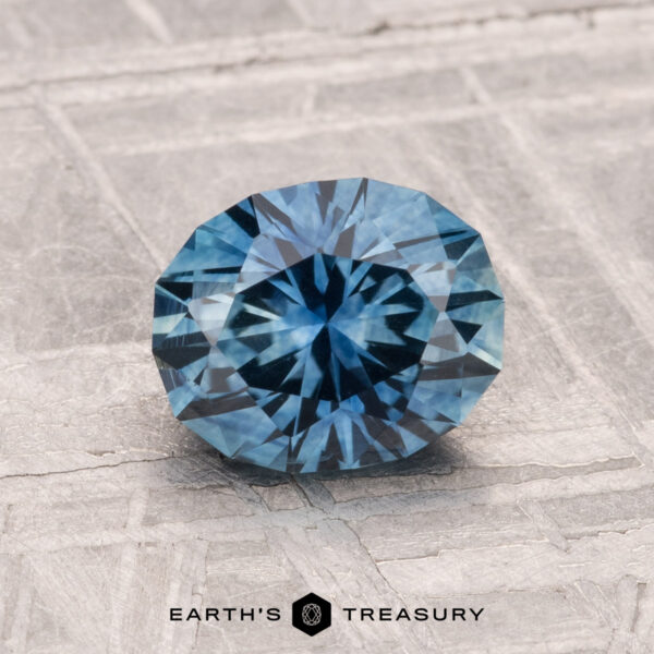 1.41-Carat Montana Sapphire (Heated) 1.41-carat heated Montana sapphire, faceted oval and blue, displayed on a textured surface with the Earth’s Treasury logo at the bottom.