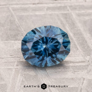 A 1.40-carat heated Montana sapphire, round and faceted, rests on a textured gray surface with "EARTH'S TREASURY" displayed below.
