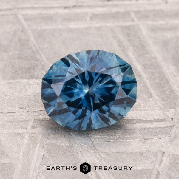 1.40-Carat Montana Sapphire (Heated) A 1.40-carat heated Montana sapphire, round and faceted, rests on a textured gray surface with "EARTH'S TREASURY" displayed below.