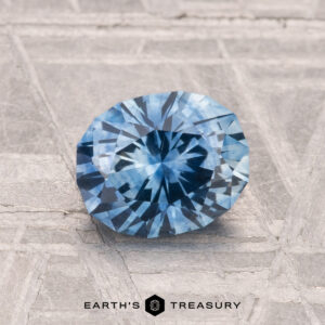 A 1.40-carat heated Montana sapphire gemstone is shown on a textured gray surface with "EARTH'S TREASURY" beneath it.
