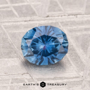 A round, faceted 1.38-carat heated Montana sapphire with a blue hue is displayed on a textured, light-colored surface with “EARTH'S TREASURY” at the bottom.