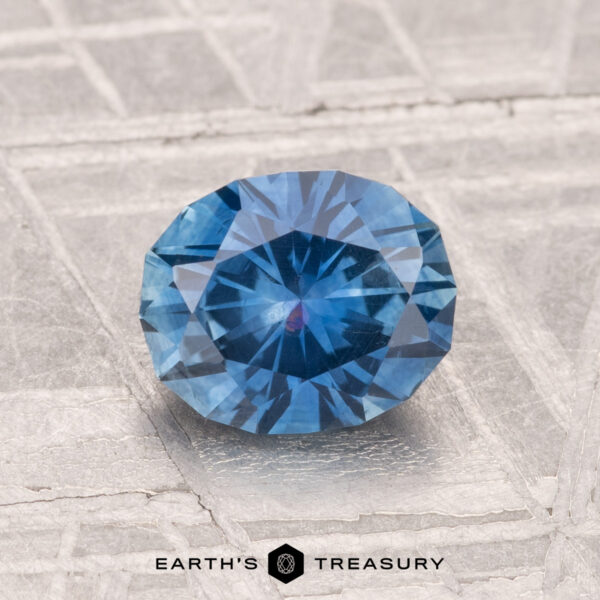 1.38-Carat Montana Sapphire (Heated) A round, faceted 1.38-carat heated Montana sapphire with a blue hue is displayed on a textured, light-colored surface with “EARTH'S TREASURY” at the bottom.