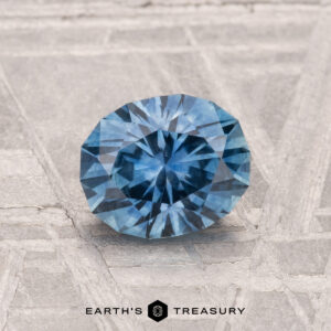 A round, faceted 1.31-carat heated Montana sapphire with a blue hue is shown on a textured gray surface, featuring the text "EARTH'S TREASURY" at the bottom.