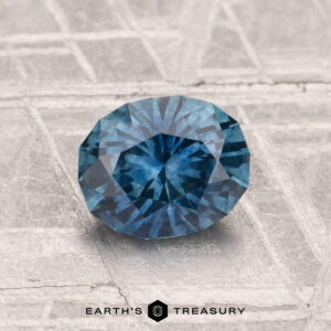 A 1.30-carat heated Montana sapphire is displayed on a textured, metallic surface with "EARTH'S TREASURY" at the bottom.