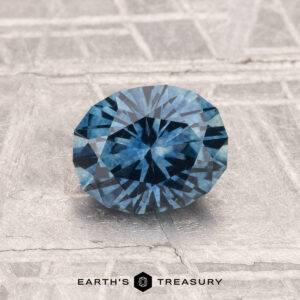 A 1.29-carat heated Montana sapphire with a blue hue rests on a gray textured surface, with "EARTH'S TREASURY" displayed at the bottom.