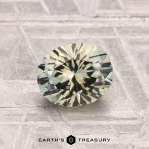 A round, faceted 1.27-carat Montana Sapphire (heated) with pale yellow and green hues rests on a textured metallic surface, with the "Earth's Treasury" logo visible at the bottom.